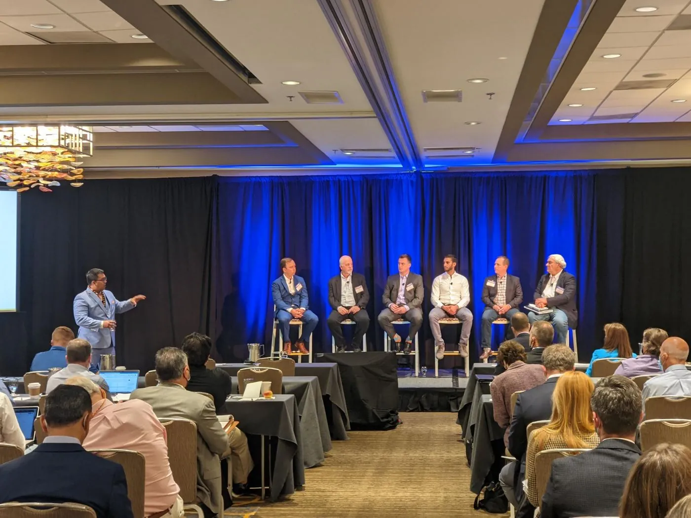 A moderator leading a six-person panel in a hotel ballroom